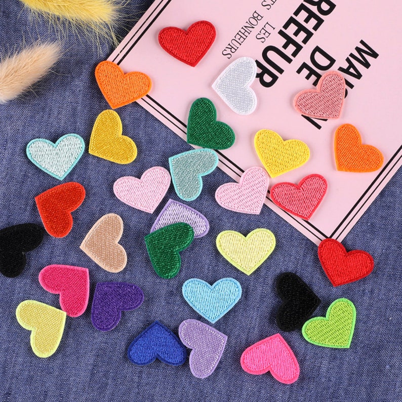 May include: Assortment of colorful felt heart patches. The patches are in various colors, including red, orange, yellow, green, blue, purple, pink, and black. The patches are arranged on a blue denim fabric background.