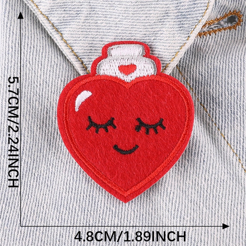 May include: Red felt heart-shaped patch with a white smiling face and closed eyes. The heart has a white top with a red heart in the center. The patch is 4.8 cm by 5.7 cm.