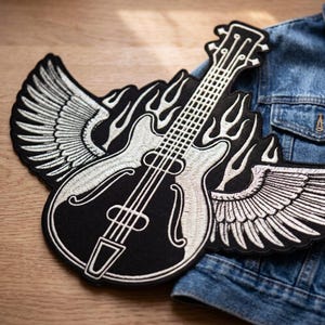 Könnte beinhalten: Schwarz-weißer, gestickter Aufnäher mit einer Gitarre, Flügeln und Flammen. Die Gitarre hat Saiten und ein Griffbrett. Die Flügel befinden sich auf beiden Seiten der Gitarre, und Flammen umgeben das Instrument. Der Aufnäher liegt auf einer Jeansjacke.