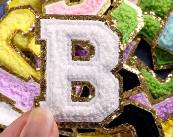 May include: A white felt letter B with gold glitter trim. The letter is surrounded by other felt letters in various colours, including yellow, pink, green, and purple.