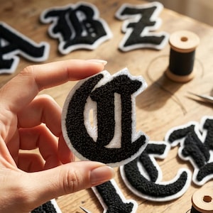 Può includere: Una toppa a forma di lettera G in bianco e nero con una superficie testurizzata e soffice e un bordo bianco. La toppa è tenuta in mano, mostrando la sua tipografia in stile gotico. Altre lettere simili e un rocchetto di filo nero sono visibili.