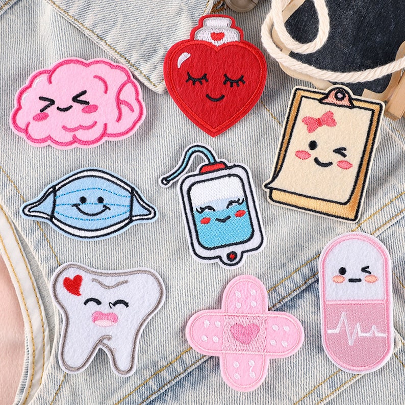 May include: Nine embroidered iron-on patches with various medical-themed designs. The patches feature a heart, brain, tooth, bandage, pill, face mask, IV bag, and a clipboard. The patches are all pink, blue, and white.