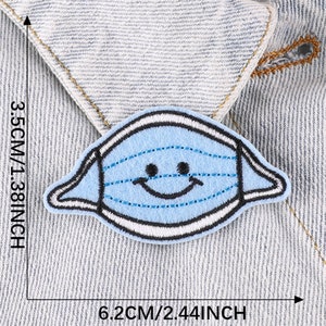 May include: Blue felt iron-on patch of a smiling face wearing a blue and white striped face mask. The patch is 6.2 cm by 3.5 cm.