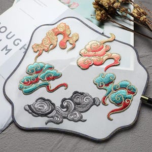 May include: A white fan with embroidered cloud designs in red, teal, gold, and grey. The fan has a dark blue border. The clouds are in various styles. The fan is on a grey surface.