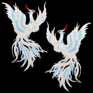 Könnte beinhalten: Zwei weiße gestickte Phönixe mit blauen und roten Akzenten. Die Phönixe sind in entgegengesetzte Richtungen gerichtet und haben lange, fließende Schwänze.