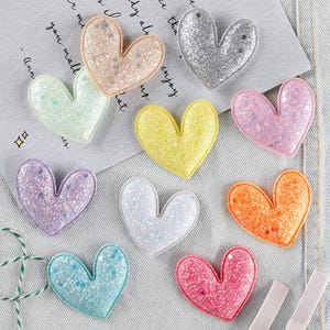 Könnte beinhalten: Eine Sammlung von neun herzförmigen Verzierungen in verschiedenen Farben, darunter Rosa, Silber, Gelb und Blau. Jedes Herz ist mit Glitzer bedeckt und hat einen genähten Rand. Diese dekorativen Herzen eignen sich für Bastelarbeiten und Verzierungen.