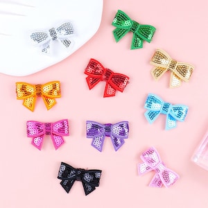 Può includere: Dieci fiocchi di paillettes colorati in vari colori, tra cui argento, verde, oro, rosso, arancione, blu, rosa, viola, nero e rosa chiaro. I fiocchi sono disposti su uno sfondo rosa.