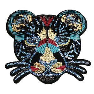 Może przedstawiać: Czarna łata z twarzy tygrysa z cekinami z niebieskimi, czerwonymi i złotymi akcentami. Oczy tygrysa są obwiedzione na niebiesko, a nos jest czerwony.