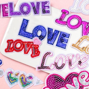 May include: Assortment of colourful iron-on patches featuring the word "LOVE" in various styles and fonts. The patches are decorated with sequins and have a shiny, glittery finish.