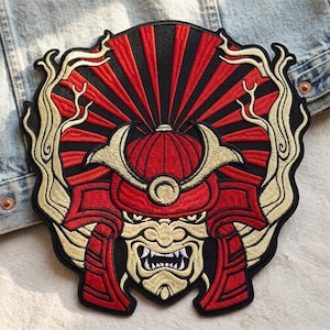 May include: Embroidered patch featuring a red and black samurai helmet design. The patch has gold accents and a detailed face with sharp teeth. The background has a sunburst pattern.