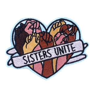 May include: An embroidered patch in the shape of a heart with five hands of different skin tones clasped together. The hands are surrounded by a light blue outline. A white banner with the text "Sisters Unite" is wrapped around the bottom of the heart.