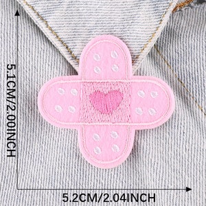 May include: Pink fabric iron-on patch shaped like a bandage with a white heart in the center. The patch is 2.04 inches wide and 2.00 inches tall.