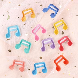 Może przedstawiać: Kolekcja kolorowych, haftowanych naszywek w kształcie nut w różnych odcieniach, w tym niebieskim, żółtym, różowym i pomarańczowym. Każda naszywka ma biały akcent i jest umieszczona na miękkim, wzorzystym tle.