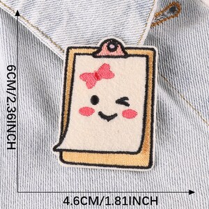 May include: A white felt patch with a yellow frame, depicting a smiling cartoon face with a pink bow. The patch is 4.6 inches wide and 2.36 inches tall.