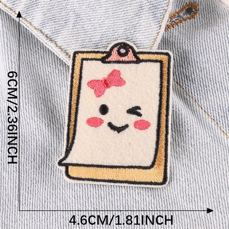 May include: A white felt patch with a yellow frame, depicting a smiling cartoon face with a pink bow. The patch is 4.6 inches wide and 2.36 inches tall.