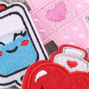 May include: Three embroidered iron-on patches. One is a red heart with a white outline and a white spray bottle on top. Another is a pink bandage with white polka dots and a pink heart in the center. The last is a blue bottle with a white outline and a smiling face with red cheeks.