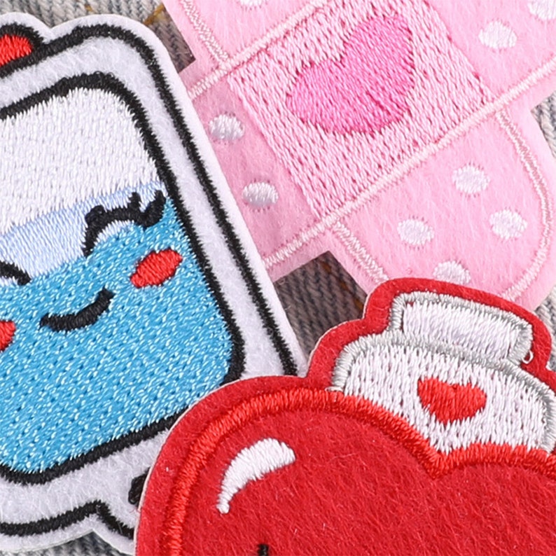 May include: Three embroidered iron-on patches. One is a red heart with a white outline and a white spray bottle on top. Another is a pink bandage with white polka dots and a pink heart in the center. The last is a blue bottle with a white outline and a smiling face with red cheeks.