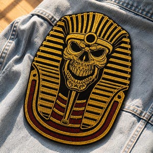 以下が含まれることがあります： ファラオの頭飾りをつけたスカルの刺繍パッチ。パッチは黒い背景に金と赤のアクセントがあります。スカルは大きく笑っており、頭飾りは縦線で詳細に描かれています。