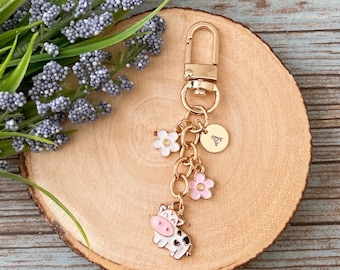 May include: Gold keychain with a cow charm, two pink flowers, a white flower, and a gold charm with the letter A.