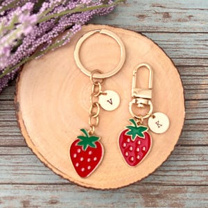 May include: Two strawberry-shaped keychains with gold-colored hardware. The strawberries are red with green tops and have a gold initial charm. One keychain has a ring, the other a clasp. The keychains rest on a wooden slice.