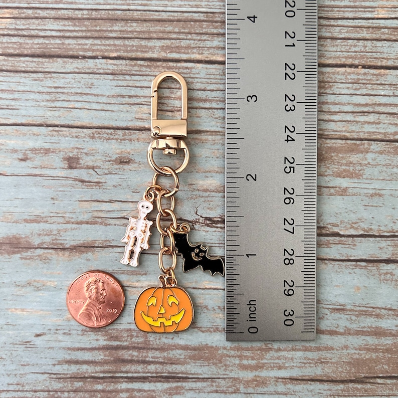 K&ouml;nnte beinhalten: Goldfarbener Halloween-Schl&uuml;sselanh&auml;nger mit einem K&uuml;rbis-, Fledermaus- und Skelett-Anh&auml;nger. Die Anh&auml;nger sind an einer goldenen Kette und einem goldenen Splitt-Ring befestigt.