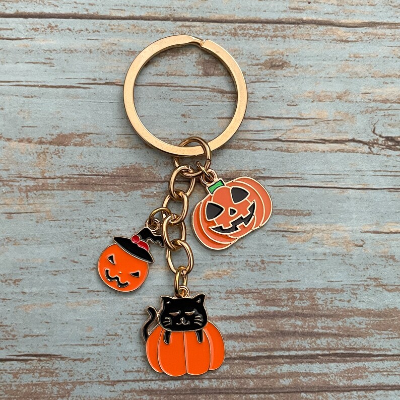 Może przedstawiać: Złoty brelok do kluczy z trzema zawieszkami w stylu Halloween: czarny kot siedzący na dyni, dynia z kapeluszem czarownicy i dynia z wyciętym uśmiechem.