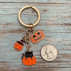 Może przedstawiać: Złoty brelok do kluczy z trzema zawieszkami w stylu Halloween: czarnym kotem z brzuchem w kształcie dyni, dynią z czarodziejskim kapeluszem i dynią z uśmiechem. Zawieszki są przymocowane do złotego łańcuszka i złotego pierścienia.