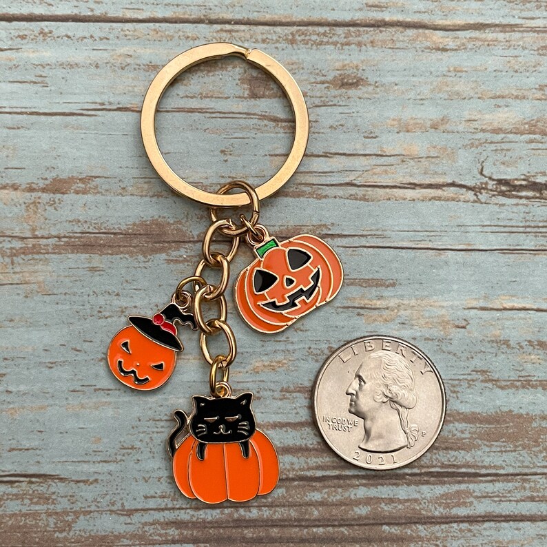 Może przedstawiać: Złoty brelok do kluczy z trzema zawieszkami w stylu Halloween: czarnym kotem z brzuchem w kształcie dyni, dynią z czarodziejskim kapeluszem i dynią z uśmiechem. Zawieszki są przymocowane do złotego łańcuszka i złotego pierścienia.