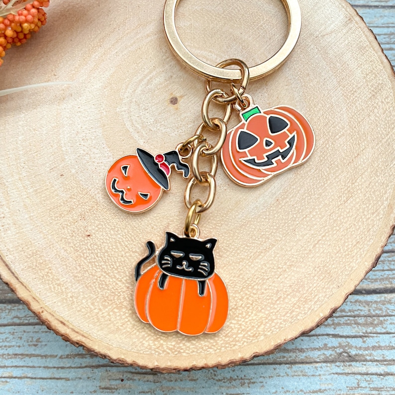 Może przedstawiać: Złoty brelok do kluczy z trzema zawieszkami w stylu Halloween: czarnym kotem siedzącym na dyni, dynią z kapeluszem czarownicy i dynią z wyciętym uśmiechem.