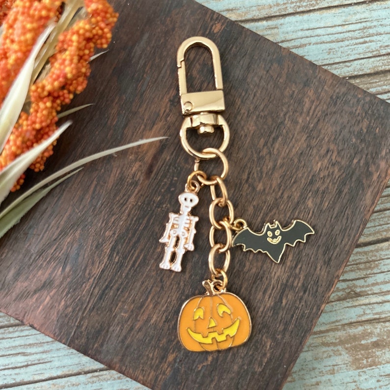 K&ouml;nnte beinhalten: Goldener Halloween-Schl&uuml;sselanh&auml;nger mit einem K&uuml;rbis-, Fledermaus- und Skelett-Anh&auml;nger.
