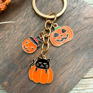 Może przedstawiać: Złoty brelok do kluczy z trzema zawieszkami w stylu Halloween: czarnym kotem siedzącym w pomarańczowej dyni, dynią z czarodziejskim kapeluszem i dynią z wyciętym uśmiechem.