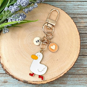 May include: A gold-toned metal keychain featuring a white duck charm with an orange beak and red feet. The keychain includes two small circular charms: one silver-toned disc engraved with the letter Z and one orange-rimmed disc with a gold heart.