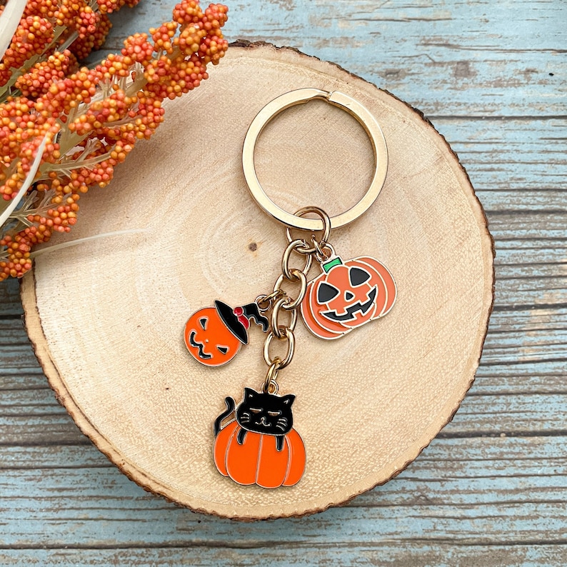 Może przedstawiać: Złoty brelok do kluczy z trzema zawieszkami w stylu Halloween: czarny kot śpiący w dyni, dynia z twarzą jack-o'-lantern i dynia z kapeluszem czarownicy.