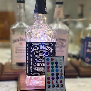 May include: A repurposed Jack Daniel's whiskey bottle with string lights inside, sitting on a wooden base. The bottle has a black label with white text and a black cap. A remote control with various color options is next to the bottle. The bottle is 10 inches tall.