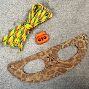 May include: A brown leather strap with a leopard print design and two holes. The strap has the text "H.I.K.E" and a small tent graphic. The strap is accompanied by a yellow and red striped shoelace and two orange plastic toggles.