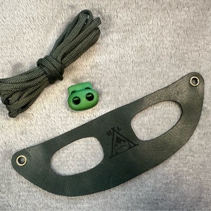 Pode incluir: Um cordão verde de paracord com um fecho de plástico verde e uma correia de couro verde escuro com dois recortes ovais e o texto "HVL" e um gráfico de tenda.
