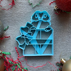 Puede incluir: Un cortador de galletas de plástico azul claro con forma de lagarto sosteniendo un regalo con un lazo. El regalo tiene una cinta y un lazo en la parte superior.