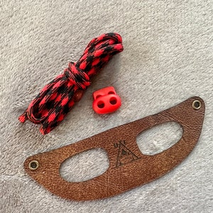 Puede incluir: Cordón de zapato rojo y negro con un cierre de cordón de plástico rojo y una correa de cuero marrón con dos agujeros y un logotipo triangular.