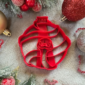 May include: Red plastic Christmas cookie cutter shaped like a snake wearing a Santa hat. The snake is curled up with its head peeking out from under the hat.