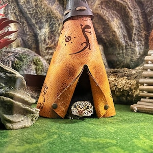 以下が含まれることがあります： 部族風のデザインが施された、小さな茶色の革製ティピー型爬虫類用隠れ家。ヒョウモントカゲモドキが入り口から顔をのぞかせています。隠れ家は緑色の表面にあり、岩の背景があります。