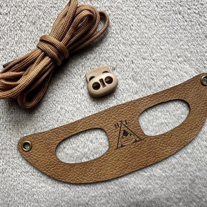 May include: A brown leather strap with two oval cutouts and a triangle logo with the text "Hike" inside. The strap is accompanied by a tan plastic toggle and a brown cord.