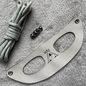 Può includere: Un cinturino in pelle bianca con due tagli ovali e il testo "HXL" in un triangolo con una volpe stilizzata all'interno. Il cinturino è accompagnato da un fermo in metallo argentato e da una corda grigia.