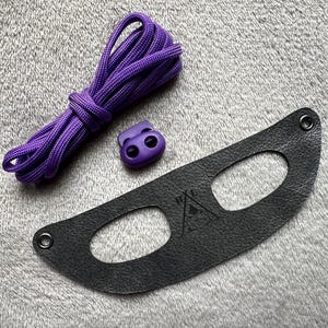 Puede incluir: Cordón de zapato morado con dos ojales de plástico morado y una correa de cuero negro con dos agujeros y un logotipo.