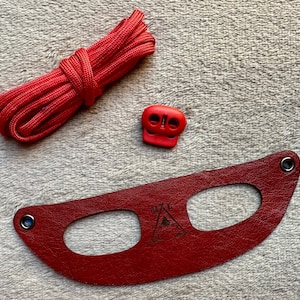Può includere: Cinturino in pelle rossa con due tagli ovali e un piccolo logo triangolare. Il cinturino è accompagnato da un cordoncino rosso e una fibbia in plastica rossa.