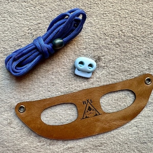 May include: A brown leather strap with two holes and the text "HXL" and a tent graphic. A blue cord and a light blue plastic toggle are also included.