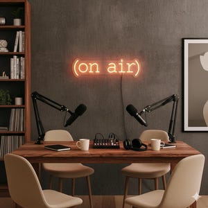 May include: A podcast studio setup featuring two microphones on adjustable arms, a mixing desk, and headphones on a wooden table. A neon sign reading "(on air)" is mounted on the wall. Beige chairs surround the table.