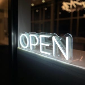 Op de afbeelding: Een wit neonbord met het woord "OPEN" op een transparante acrylbasis. Het bord is verlicht, de letters gloeien helder. Het bevindt zich in een raam met een vage achtergrond.