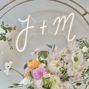 Può includere: Un'insegna al neon con le lettere "J + M" in un carattere decorativo, sullo sfondo di una composizione floreale. I fiori includono fiori bianchi, rosa e arancioni, con accenti verdi. L'insegna è illuminata da una luce soffusa e calda.