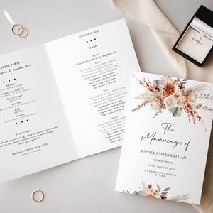 May include: A wedding stationery set comprising a tri-fold order of service and an invitation. The invitation is adorned with floral designs in brown, beige, and green. Gold wedding rings and a ring box are also visible.