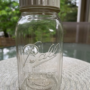 May include: A clear glass Ball Perfect Mason jar with a cream-colored lid. The jar has the Ball logo and "Perfect Mason" embossed on the front. The jar is sitting on a light-colored, woven placemat.